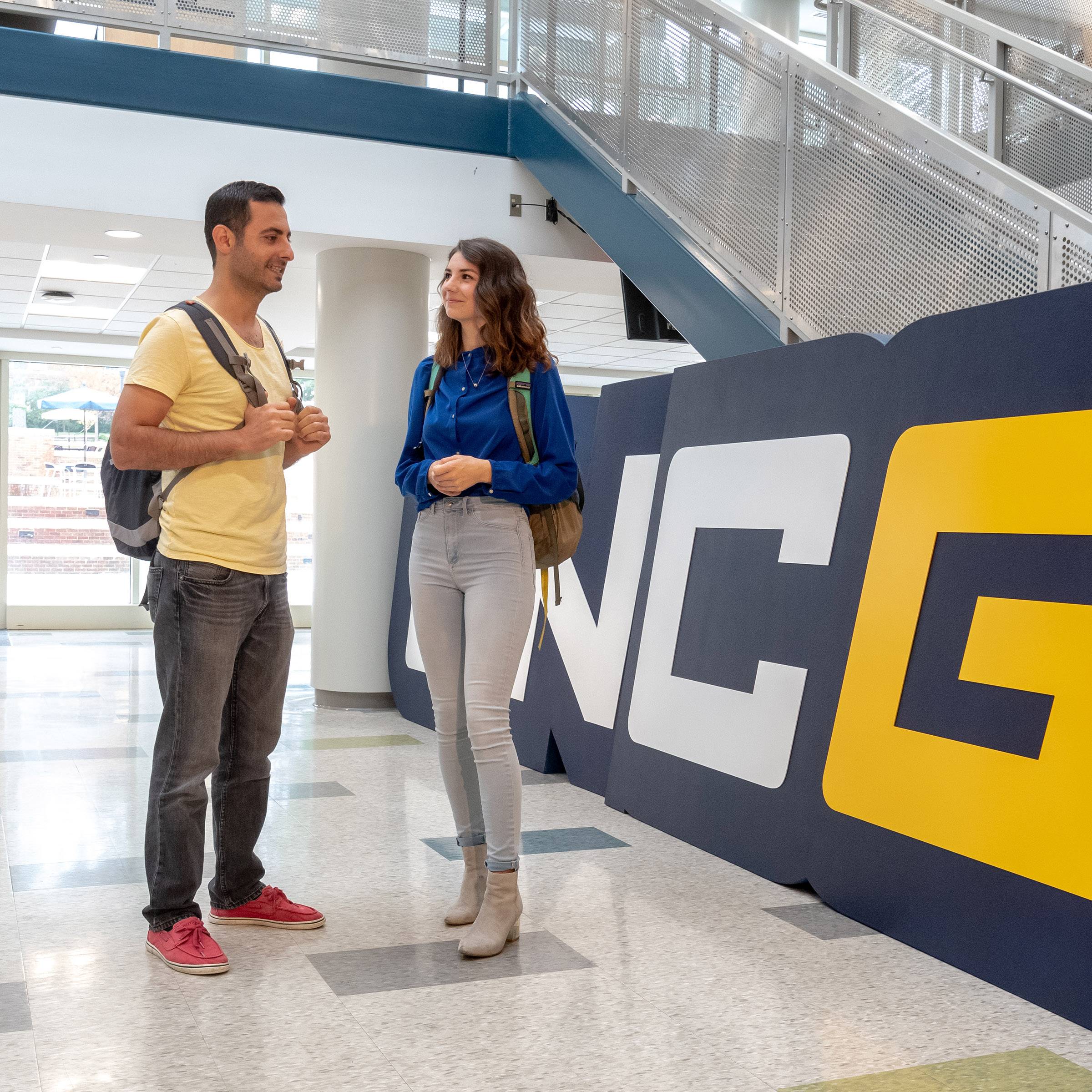 Two students talk in front of the UNCG sign inside Moran Commons.